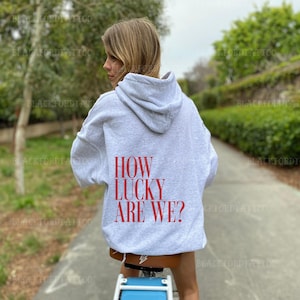 以下が含まれることがあります： 背面に赤い文字で「HOW LUCKY ARE WE?」とプリントされた灰色のフード付きスウェットシャツです。