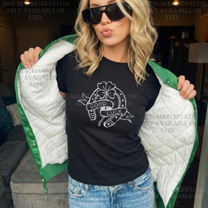 May include: Black t-shirt with a white "Good Luck" horseshoe and clover design. The person is wearing a green satin jacket with a white quilted lining and blue jeans. The text "SHOP BLKFORDTATTOO ONLY AVAILABLE ON ETSY" is visible.