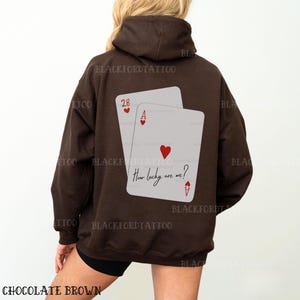 Puede incluir: Una sudadera con capucha marr&oacute;n con un gr&aacute;fico de dos cartas de juego en la espalda. Las cartas son un as de corazones y un 28 de corazones. El texto "How lucky are we?" est&aacute; escrito en el as de corazones.