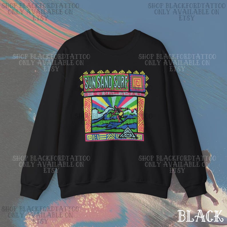 May include: Black sweatshirt with a colorful graphic featuring the text "SUN SAND SURF" above a surfer riding a wave. The design includes a rainbow, an eye symbol, and geometric patterns. The sweatshirt is a casual wear item.