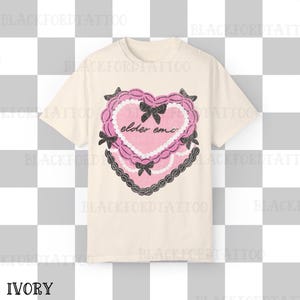 May include: Ivory-colored t-shirt featuring a pink heart design with the text "elder emo." The heart is decorated with black bows and a black lace border. The shirt is a short-sleeved crew neck.