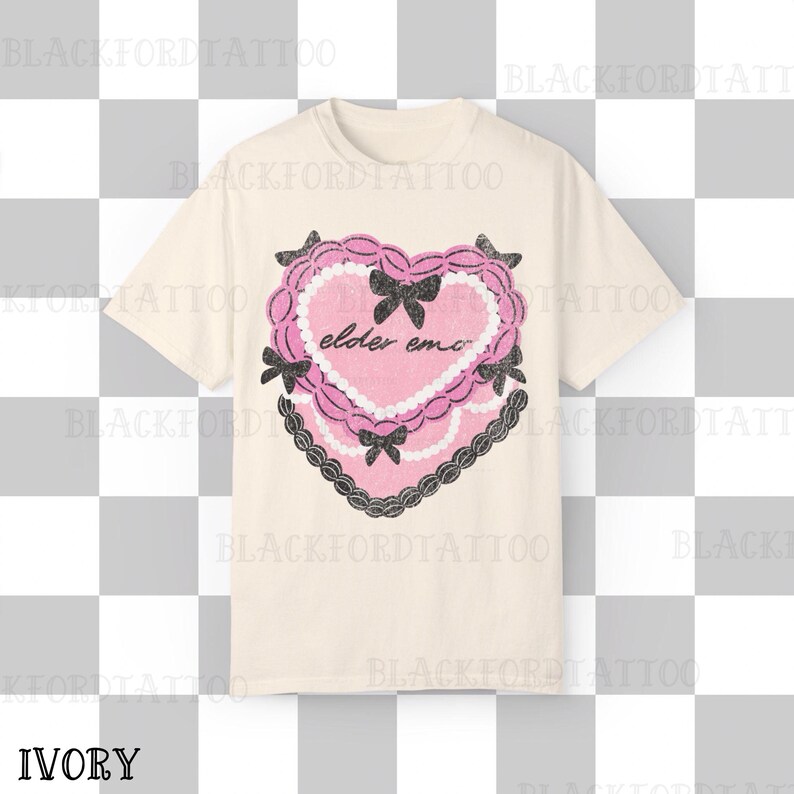 May include: Ivory-colored t-shirt featuring a pink heart design with the text "elder emo." The heart is decorated with black bows and a black lace border. The shirt is a short-sleeved crew neck.
