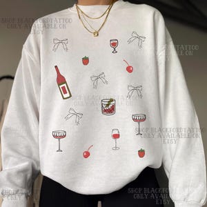 Op de afbeelding: Witte sweatshirt met een patroon van wijnglazen, strikken, kersen, aardbeien en een wijnfles in zwart en rood. De tekst "SHOP BLACKFORDTATTOO ONLY AVAILABLE ON ETSY" is op het sweatshirt gedrukt.