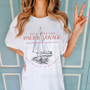 Peut inclure: T-shirt blanc avec un graphique d'un voilier sur l'océan. Le texte sur le t-shirt est "Deep Blue Sea Pacific Voyage Original Spirit Pacific Soul The Ocean Is My Home".