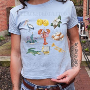 May include: A light blue t-shirt with a variety of New Orleans themed illustrations, including a pelican, crawfish, fleur-de-lis, beignets, corn on the cob, a trumpet, a glass of whiskey, and an alligator. The text "Blackford Tattoo" is visible on the shirt.