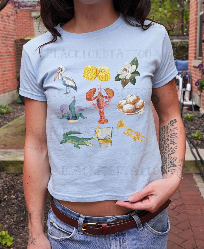May include: A light blue t-shirt with a variety of New Orleans themed illustrations, including a pelican, crawfish, fleur-de-lis, beignets, corn on the cob, a trumpet, a glass of whiskey, and an alligator. The text "Blackford Tattoo" is visible on the shirt.