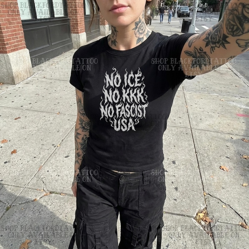 May include: Black t-shirt with the text "NO ICE NO KKK NO FASCIST USA" in a flame-like design. The shirt is short-sleeved and worn with black cargo pants. The image is taken outdoors on a sunny day.