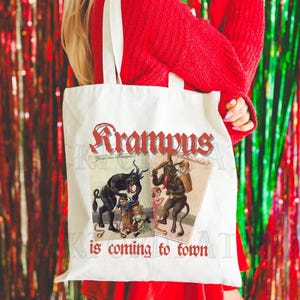 May include: A white tote bag with the text "Krampus is coming to town" in red. The bag features two vintage illustrations of Krampus, a horned figure from European folklore, with the text "Gruss vom Krampus" and "B&ouml;sewichte!" in German.