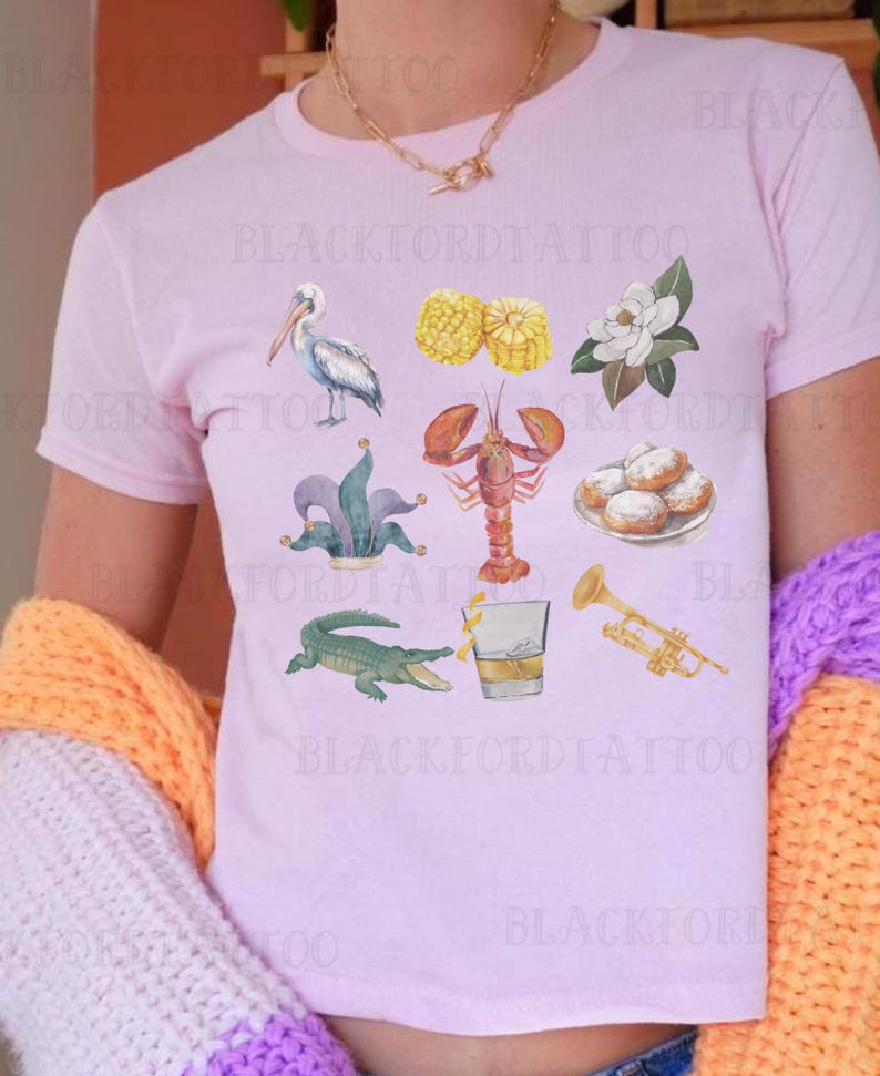 May include: A pink t-shirt with a variety of colorful illustrations of New Orleans themed imagery, including a pelican, a crawfish, a fleur-de-lis, a trumpet, a glass of whiskey, beignets, and corn on the cob.