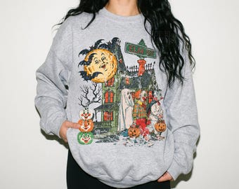 May include: A heather grey crewneck sweatshirt featuring a graphic print of a haunted house, a large yellow moon with a face, and a stack of three jack-o'-lanterns. The design includes a sign reading Elm St and figures dressed in Halloween costumes.