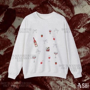 May include: A light gray sweatshirt with a repeating pattern of wine glasses, cherries, strawberries, bows, and a wine bottle. The text "SHOP BLACKFORDTATTOO ONLY AVAILABLE ON ETSY" is printed on the sweatshirt.