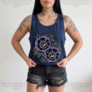 May include: Navy blue tank top featuring a large graphic of two purple roses with green and orange leaves. The text "SHOP BLACKFORDTATTOO ONLY AVAILABLE ON ETSY" is printed on the top and bottom of the image. The tank top is paired with black denim shorts.