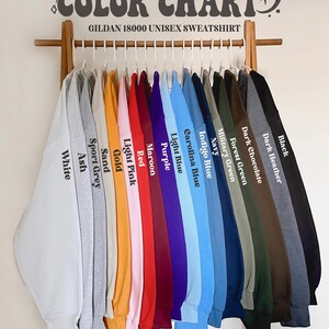 May include: A color chart displaying a variety of Gildan 18000 unisex sweatshirts. The sweatshirts are arranged on a wooden rack, showcasing colors like white, ash, sport grey, sand, gold, light pink, red, maroon, purple, light blue, carolina blue, indigo blue, navy, military green, forest green, dark chocolate, dark heather, and black.