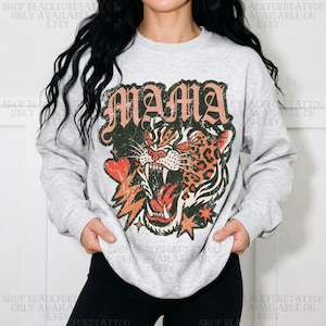 May include: A light gray crewneck sweatshirt featuring a large graphic print of a roaring leopard head with a distressed texture. The word MAMA is printed in a bold, vintage-style font above the leopard, accompanied by a red heart and star icons.