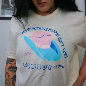 May include: A white t-shirt with a blue and pink cowboy hat graphic and the text "Misgendering people isn't very cowboy of you".