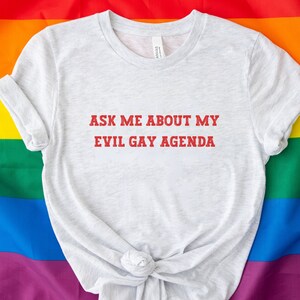 May include: A light gray t-shirt with the text "ASK ME ABOUT MY EVIL GAY AGENDA" in red letters. The shirt is displayed on a rainbow flag background, representing the LGBTQ+ community.