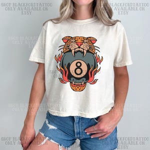 May include: Cream-colored t-shirt featuring a graphic of a tiger head biting an eight ball surrounded by flames. The design is in shades of orange, red, and black. The text "SHOP BLACKFORDTATTOO ONLY AVAILABLE ON ETSY" is printed around the image.