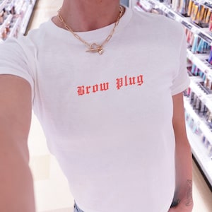 May include: White t-shirt with a red "Brow Plug" graphic in a gothic font. The shirt is worn by a person with a gold chain necklace.