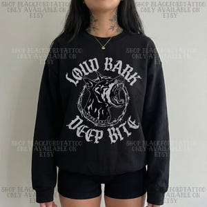 以下が含まれることがあります： 黒いスウェットシャツ。白いゴシック体で「LOUD BARK DEEP BITE」と書かれています。デザインは、有刺鉄線の円の中に、うなり声を上げている犬の頭が描かれています。クルーネックのスタイルです。