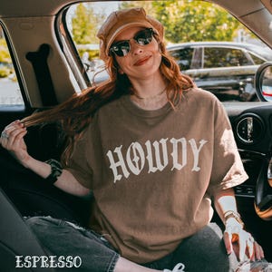May include: A woman wearing a brown t-shirt with the word "HOWDY" printed in white. She is sitting in a car and has her hair pulled back.