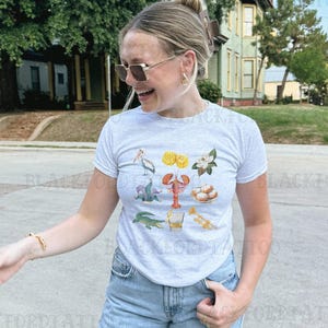 May include: A gray t-shirt with a graphic print of various New Orleans themed icons, including a pelican, a crawfish, a fleur-de-lis, beignets, a glass of whiskey, and a trumpet.
