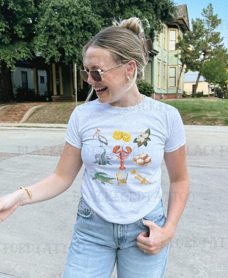 May include: A gray t-shirt with a graphic print of various New Orleans themed icons, including a pelican, a crawfish, a fleur-de-lis, beignets, a glass of whiskey, and a trumpet.