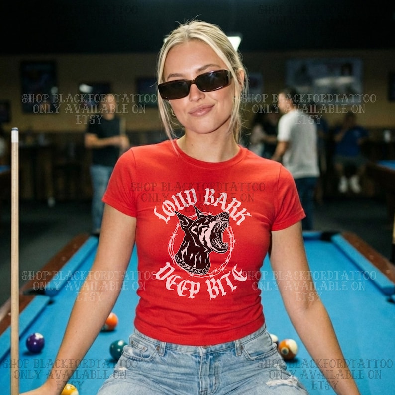 Puede incluir: Camiseta roja de cuello redondo con un gr&aacute;fico de un perro gru&ntilde;endo y las palabras "Loud Bark Deep Bite" en blanco. La camiseta se usa con vaqueros azules. La imagen est&aacute; tomada en un sal&oacute;n de billar.