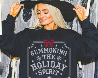 Op de afbeelding: Een zwarte sweatshirt met de tekst "SUMMONING THE HOLIDAY SPIRIT" in een decoratief kader, met een rode strik en een planchette-afbeelding. Onderdeel van een Halloween- of vakantie-thema collectie.
