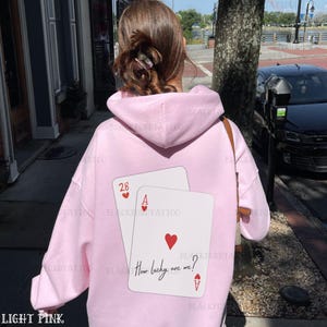Puede incluir: Una sudadera con capucha rosa claro con un gr&aacute;fico de dos cartas de juego, un as y un 28, con corazones. El texto "How lucky are we?" est&aacute; impreso en las cartas.