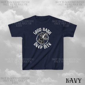 Puede incluir: Camiseta azul marino con un gr&aacute;fico en blanco y negro de la cabeza de un perro gru&ntilde;endo, rodeada de alambre de p&uacute;as, con las palabras "LOUD BARK DEEP BITE" en blanco. La palabra "NAVY" est&aacute; en la parte inferior derecha. El nombre de la tienda Etsy tambi&eacute;n est&aacute; impreso.