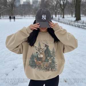 May include: A beige sweatshirt featuring a deer illustration and the text "BLACKFORDTATTOO AVAILABLE ON ETSY". The person is wearing a grey baseball cap with a white "NY" logo. The background is a snowy park.