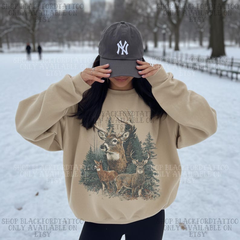 May include: A beige sweatshirt featuring a deer illustration and the text "BLACKFORDTATTOO AVAILABLE ON ETSY". The person is wearing a grey baseball cap with a white "NY" logo. The background is a snowy park.
