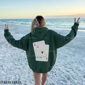 Puede incluir: Una sudadera con capucha de color verde bosque con un gr&aacute;fico de dos cartas de juego en la espalda. Las cartas son un 28 y un as de corazones. El texto en el as de corazones dice "How lucky are we?"