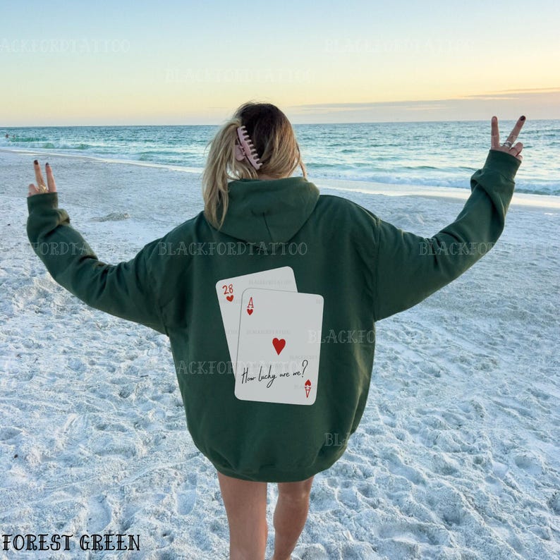Puede incluir: Una sudadera con capucha de color verde bosque con un gr&aacute;fico de dos cartas de juego en la espalda. Las cartas son un 28 y un as de corazones. El texto en el as de corazones dice "How lucky are we?"