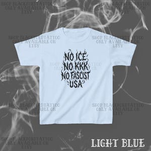May include: Light blue t-shirt with the text "NO ICE, NO KKK, NO FASCIST, USA" in a stylized black font. The shirt has short sleeves and a crew neck. The Etsy shop name is printed around the shirt.