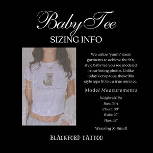 May include: A white cropped t-shirt with a cartoon cat graphic and the text "I'm literally just a girl". The model is wearing a size X-Small. The text "Baby Tee Sizing Info" is at the top of the image. The text "Blackford Tattoo" is at the bottom of the image. The model is 5'7" and weighs 120 lbs. Her bust is 34A, chest is 33 inches, waist is 27 inches, and hips are 32 inches.