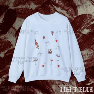 May include: Light blue sweatshirt with a pattern of wine bottles, glasses, cherries, strawberries, and bows. The text "SHOP BLACKFORDTATTOO ONLY AVAILABLE ON ETSY" is printed on the sweatshirt.