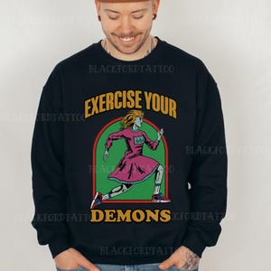 Könnte beinhalten: Ein schwarzes Sweatshirt mit einem Grafikdruck einer laufenden Frau mit dem Text "Exercise Your Demons" und der Nummer "666" auf ihrem Shirt.