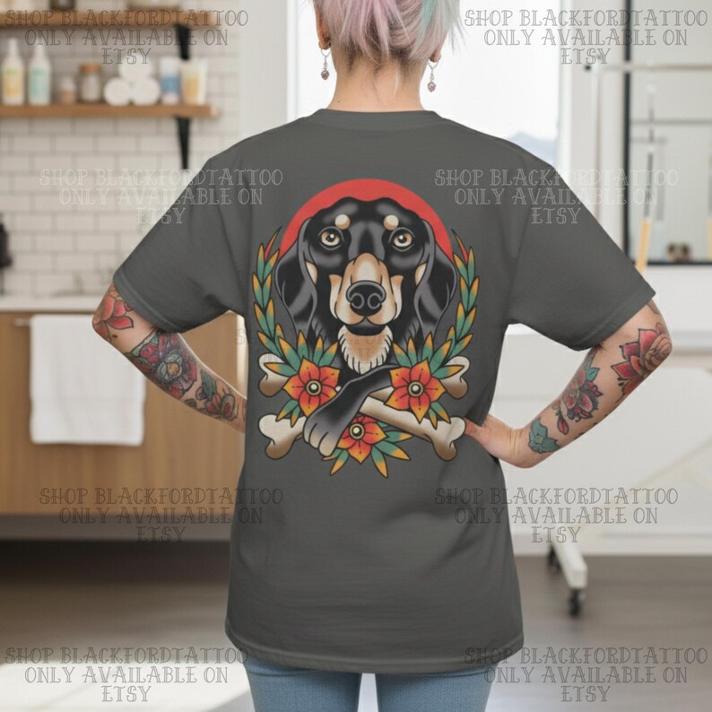 K&ouml;nnte beinhalten: Dunkelgraues T-Shirt mit einer Illustration eines Dackelhundes, einem roten Heiligenschein, Lorbeerkranz, Blumen und gekreuzten Knochen. Der Text "SHOP BLACKFORDTATTOO ONLY AVAILABLE ON ETSY" ist um das Bild gedruckt.