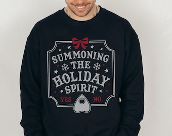 Op de afbeelding: Een zwart sweatshirt met een wit grafisch ontwerp met de tekst "Summoning the Holiday Spirit" met een rode strik en een Ouija-bord afbeelding. De tekst "Ja" en "Nee" staat onder het bord.