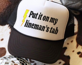Op de afbeelding: Een zwart-witte truckercap met een zwart mesh-achterpaneel. De voorkant van de cap heeft een gele bliksem en de tekst "Put it on my lineman's tab".