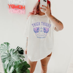 May include: White oversized t-shirt with a graphic of two pink bear heads with a heart and dagger in between. The text "Thick Thighs Sleep Deprived Muscle Mommy Lifting Club" is printed below the bears.