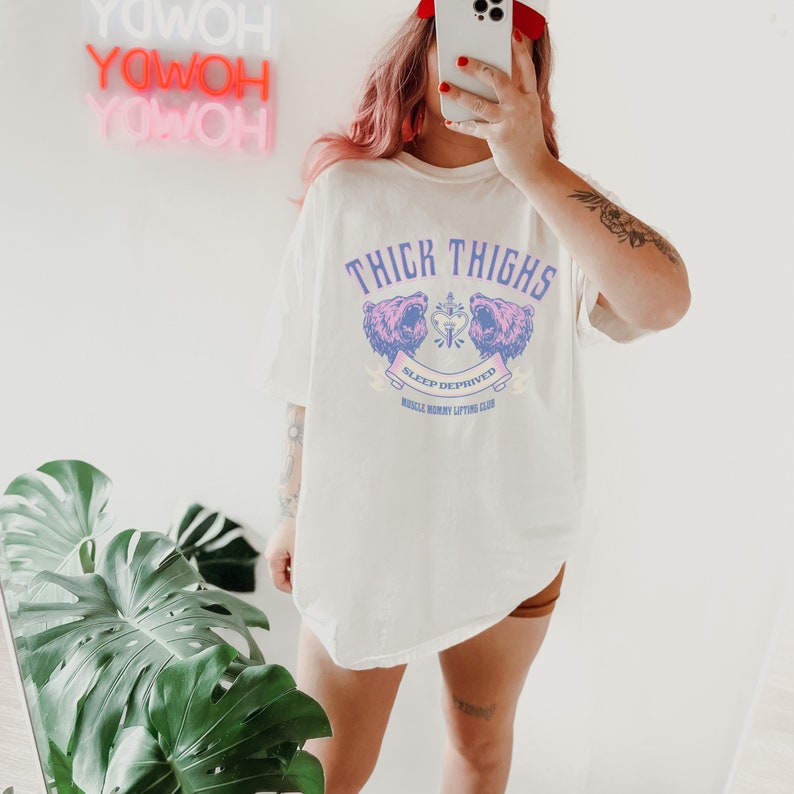 May include: White oversized t-shirt with a graphic of two pink bear heads with a heart and dagger in between. The text "Thick Thighs Sleep Deprived Muscle Mommy Lifting Club" is printed below the bears.