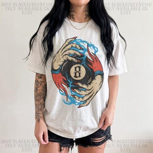 May include: Cream-colored t-shirt featuring a graphic of a black 8-ball being held by two hands with red and blue accents. The hands have long red fingernails. The text "SHOP BLACKFORDTATTOO ONLY AVAILABLE ON ETSY" is printed on the top and bottom of the shirt.
