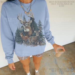 May include: A pale blue sweatshirt with a detailed graphic of deer in a forest scene. The design includes three deer, trees, and a snowy ground. The text "SHOP BLACKFORDTATTOO ONLY AVAILABLE ON ETSY" is printed around the sweatshirt.