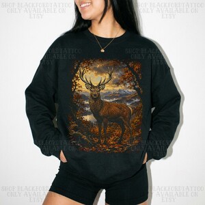 May include: A black long-sleeve sweatshirt featuring a large rectangular graphic print on the front. The graphic depicts a brown stag standing in a forest clearing with autumn foliage, mountains in the distance, and a golden sunset sky.