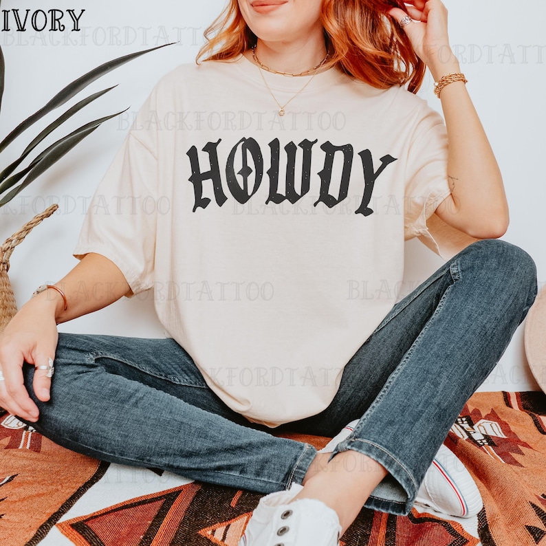 May include: A woman wearing a light beige t-shirt with the word "Howdy" printed in black gothic lettering. She is wearing blue jeans and white sneakers.