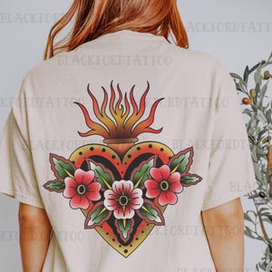 May include: A beige t-shirt with a traditional tattoo style image of a flaming heart with flowers. The heart is red, yellow, and black, and the flowers are red and green.