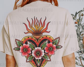 Könnte beinhalten: Ein beiges T-Shirt mit einem traditionellen Tattoo-Motiv eines brennenden Herzens mit Blumen. Das Herz ist rot, gelb und schwarz, und die Blumen sind rot und grün.