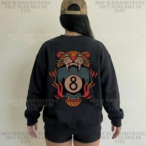 May include: Black sweatshirt featuring a vibrant graphic of a snarling leopard above a flaming eight-ball. The design incorporates orange, red, and teal colors. The text "SHOP BLACKFORDTATTOO ONLY AVAILABLE ON ETSY" is printed on the top and bottom of the image.